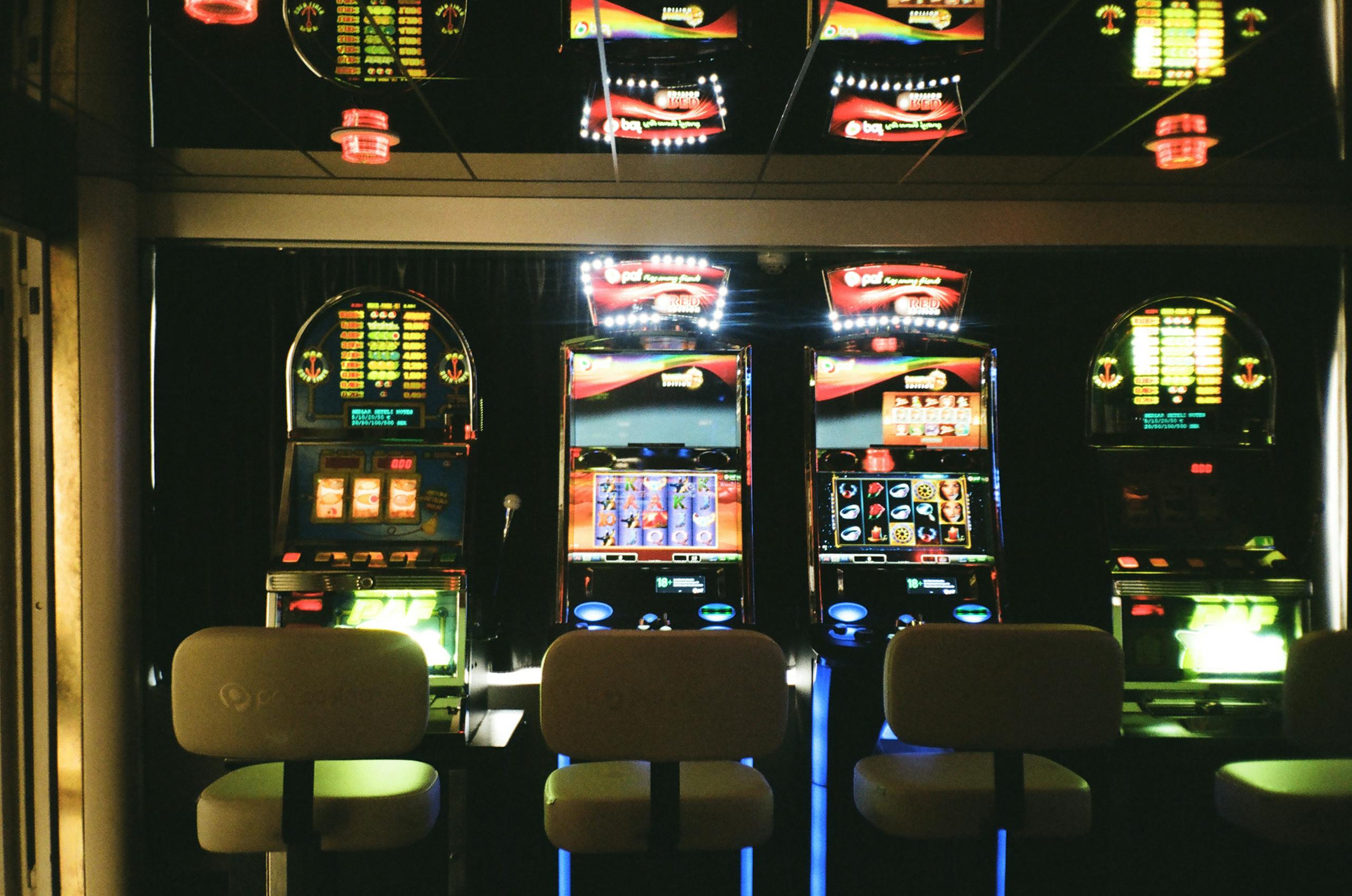 Slot machines in a dimly lit casino with reflective ceiling and empty chairs.